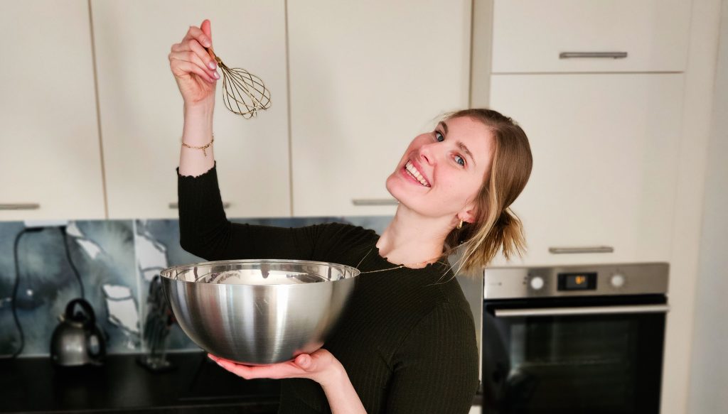 Lachende Frau mit blonden Haaren hält eine Rührschüssel und einen Schneebesen