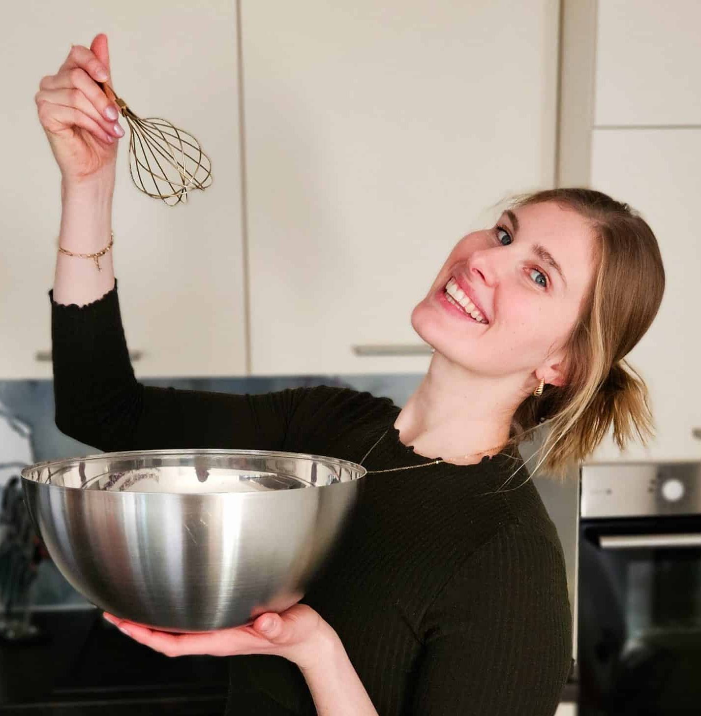 Lachende Frau mit blonden Haaren hält eine Rührschüssel und einen Schneebesen