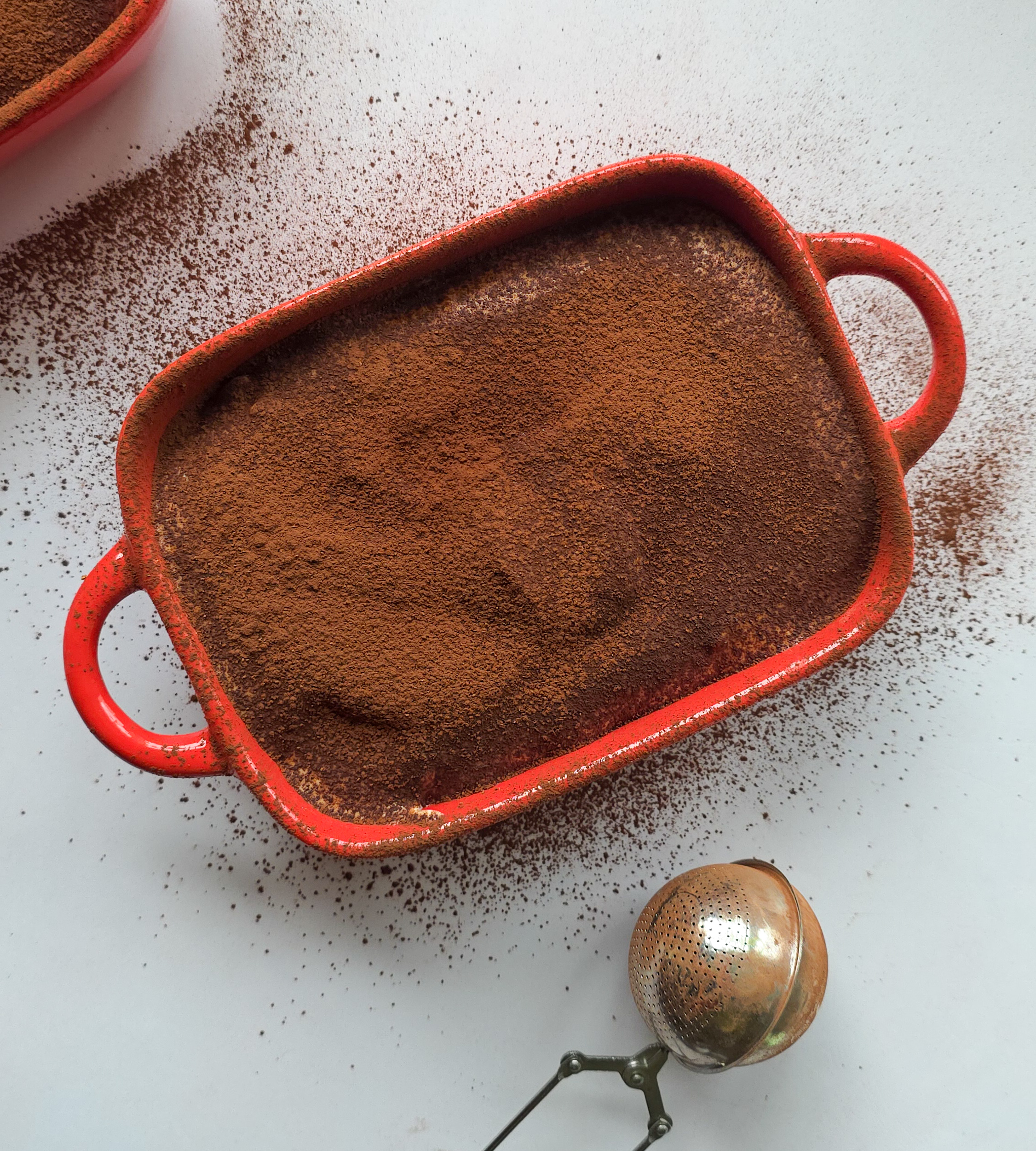 Veganes proteinreiches Baked Oats Dessert in roter Auflaufform, bestreut mit Kakaopulver, goldene Teesieb daneben.