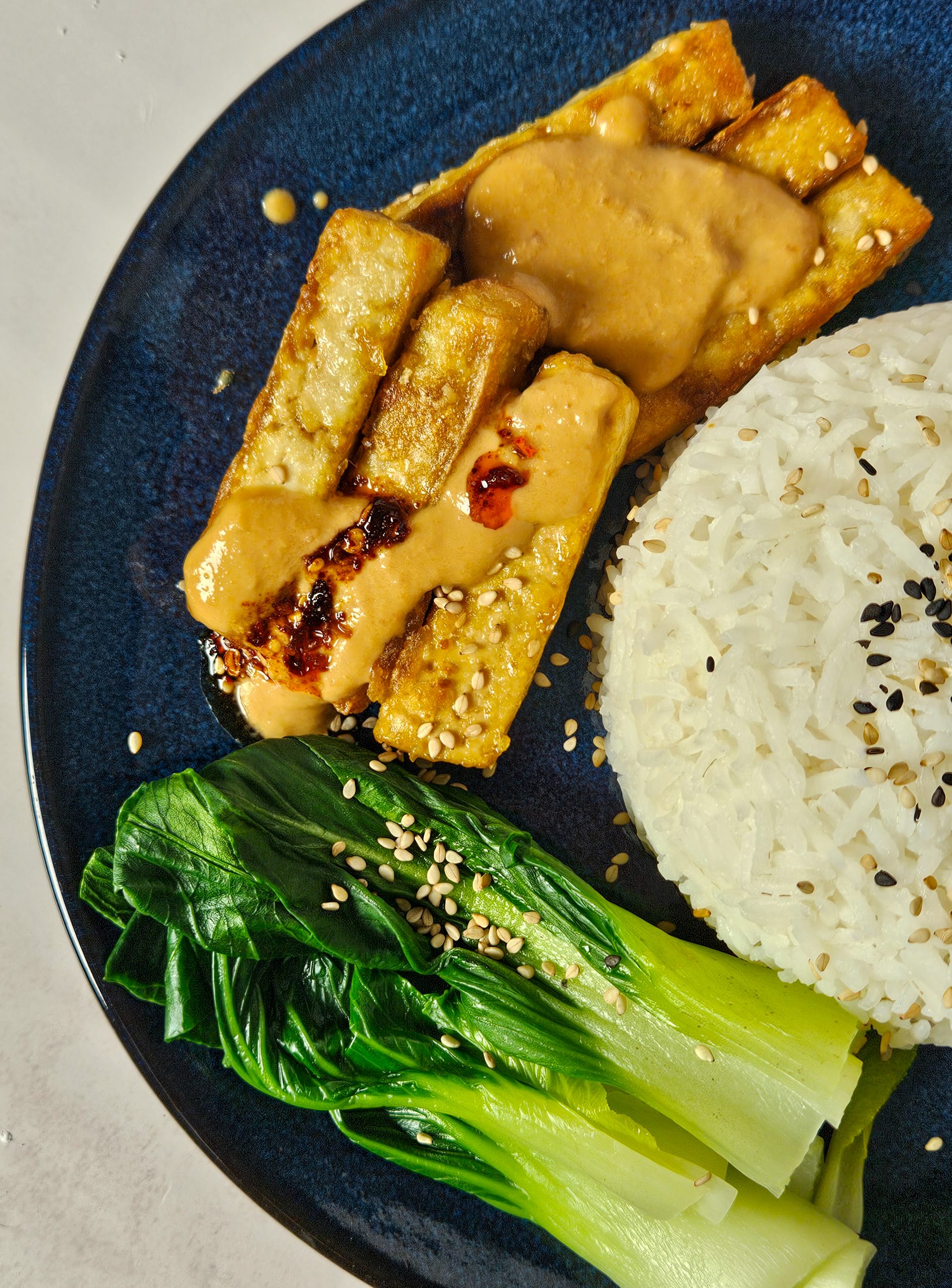 Knusprig angebratene Tofu-Streifen mit Erdnusssoße und Chiliöl, dazu Reis, Pak Choi und Sesam.