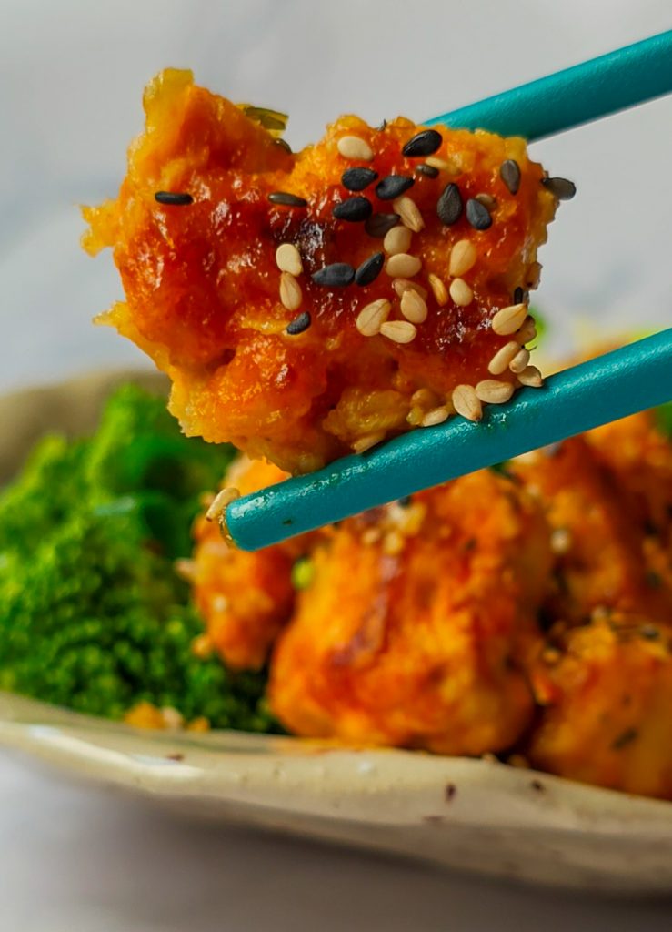 Mit Panade und Sesam ummantelter Tofu zwischen zwei Ess-Stäbchen vor einem Teller mit Tofu und Brokkoli.