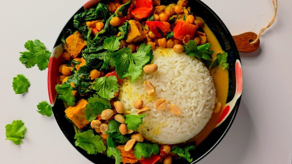 Bunte vegane Curry-Bowl mit weißem Reis, geröstetem Kürbis, Kichererbsen, Spinat, Paprika, Erdnüssen und Koriander.