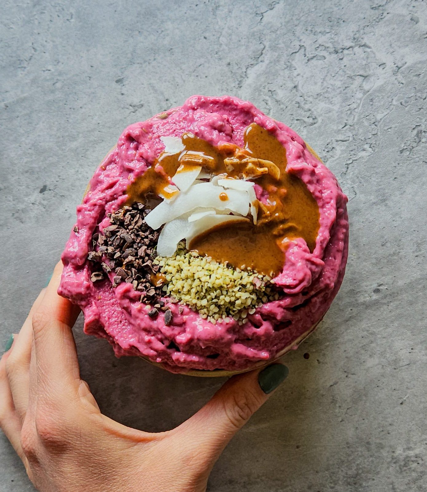 Eine Hand hält eine pinke gefrorene Smoothiebowl in einer Schale, die mit Kakaonibs, Hanfsamen, Kokoschips und Mandelmus getoppt ist