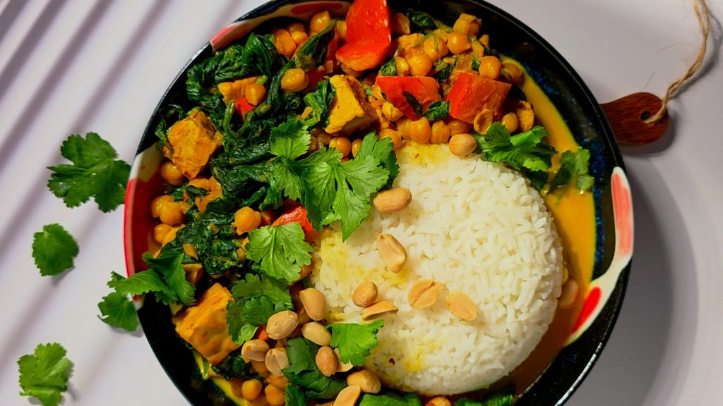 One-Pot Kürbis Curry: buntes Gemüse, Kichererbsen und Tempeh in cremiger Sauce, angerichtet mit Reis, frischem Koriander und Erdnüssen.