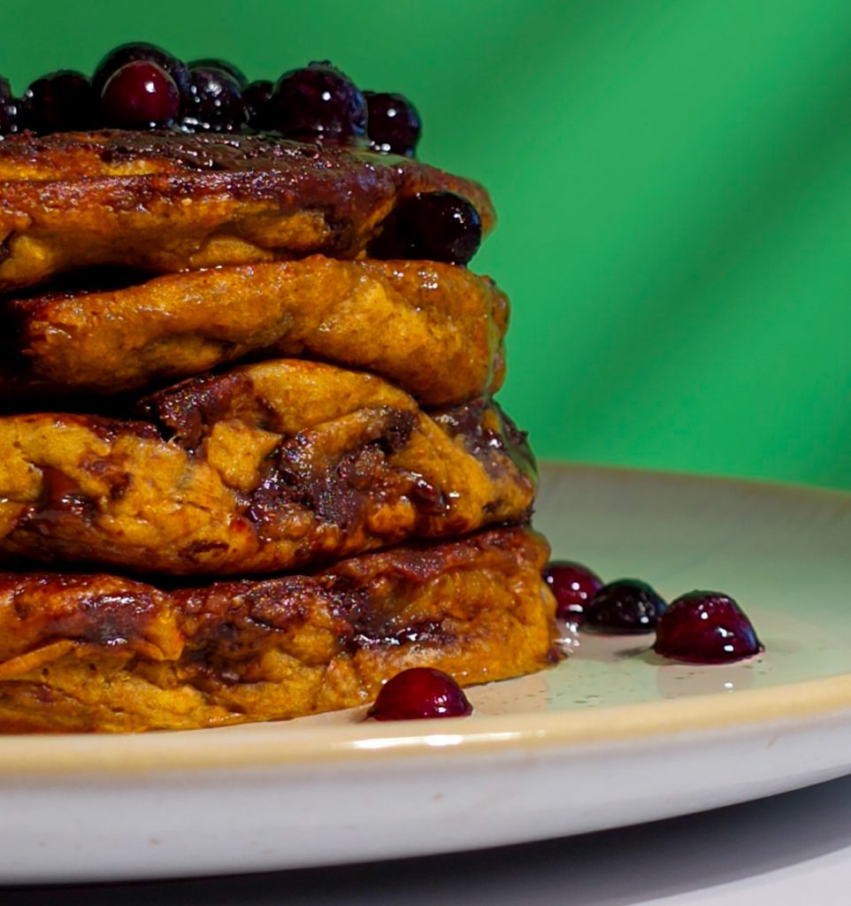 Fluffige vegane Pancakes mit Schokostückchen, auf einem hellen Teller serviert, vor leuchtend grünem Hintergrund.