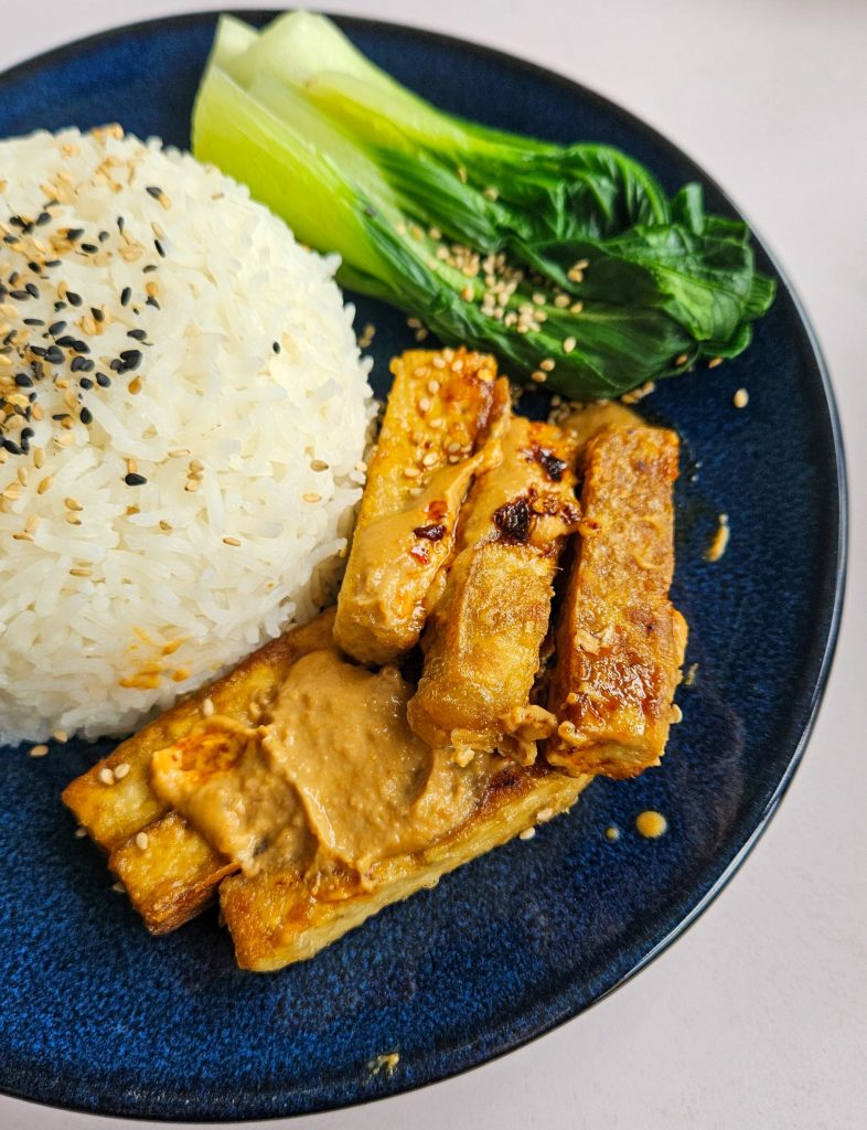 Knusprig angebratene Tofu-Streifen mit Erdnusssoße und Chiliöl, dazu Reis, Pak Choi und Sesam.