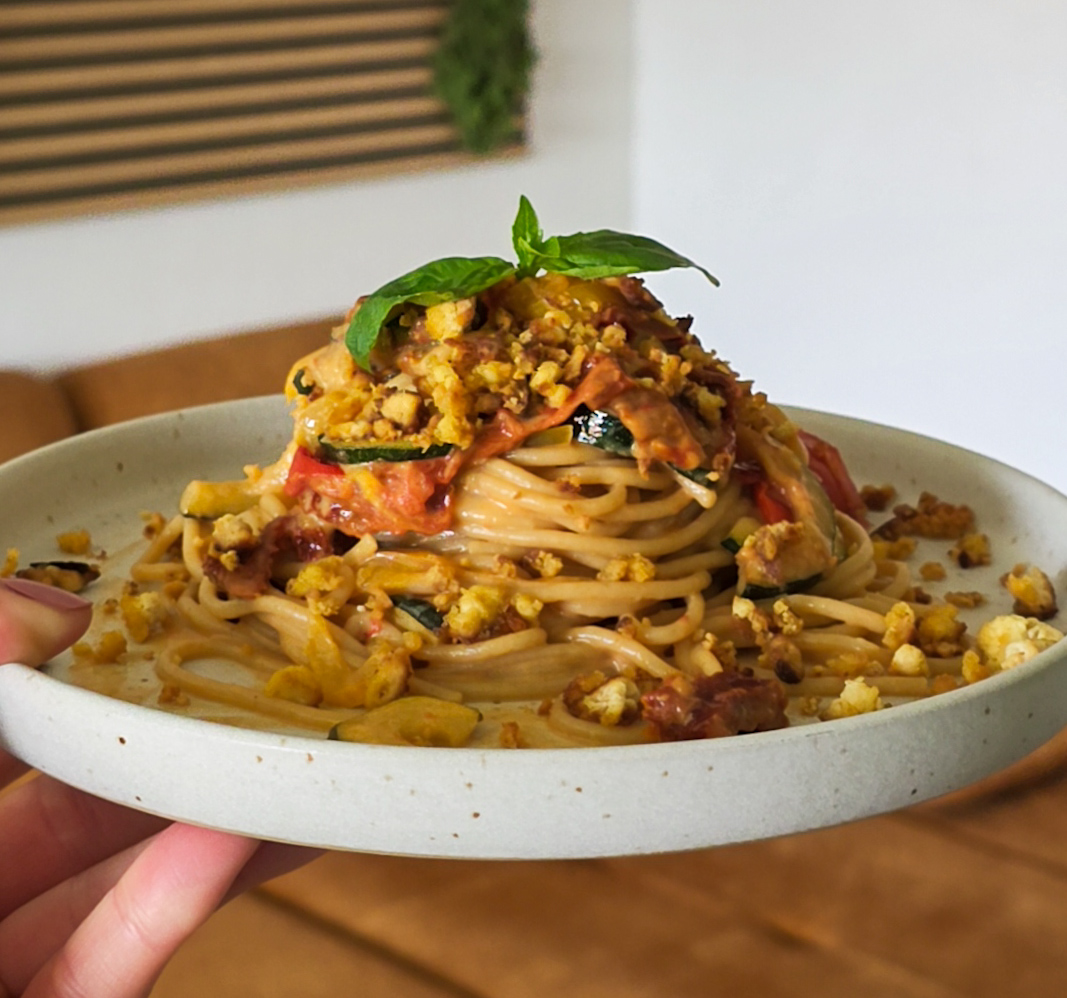 Seitliche Nahaufnahme eines Bergs Spaghetti mit gebratener gelber Paprika, Zucchini und Tomaten. Knruspriger Tofu und frisches Basilikum liegen obenauf.