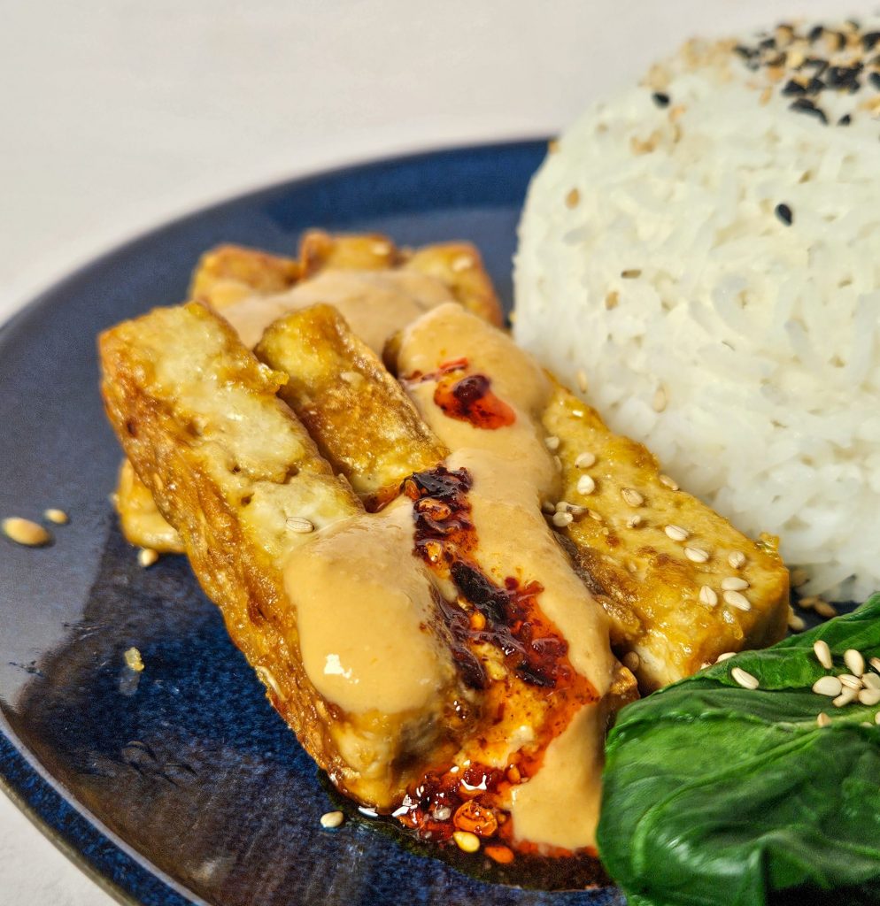 Knusprig angebratene Tofu-Streifen mit Erdnusssoße und Chiliöl, dazu Reis, Pak Choi und Sesam.