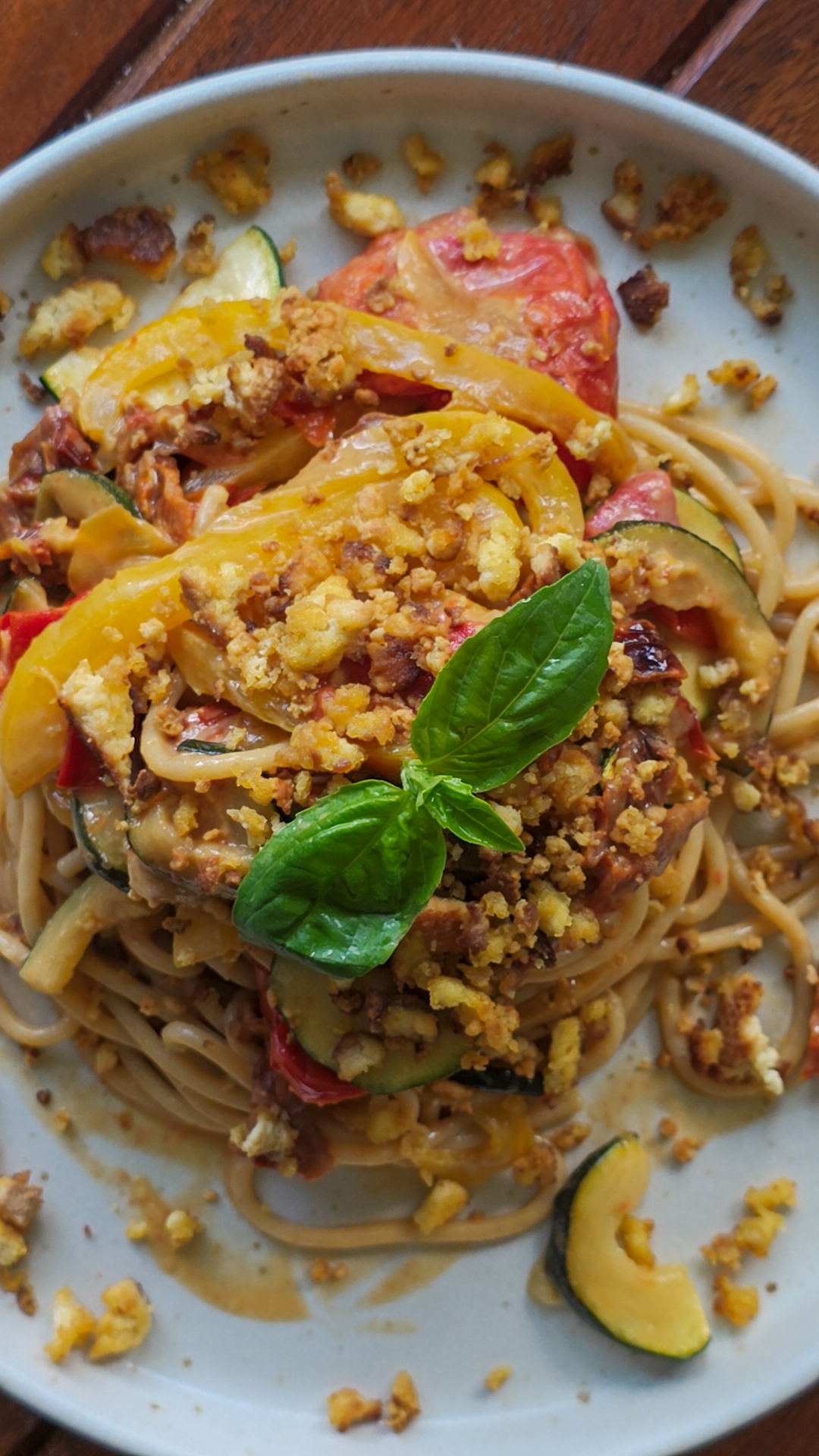 Tofu zubereiten: Draufsicht auf einen Teller Spaghetti mit gebratenem Gemüse, darunter gelbe Paprika, Zucchini und Tomaten. Das Gericht ist mit knusprigen veganen Bröseln und frischen Basilikumblättern garniert.