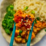 Mit Panade und Sesam ummantelter knuspriger Tofu zwischen zwei Ess-Stäbchen über einem Teller mit Tofu, Mie-Nudeln und Brokkoli.