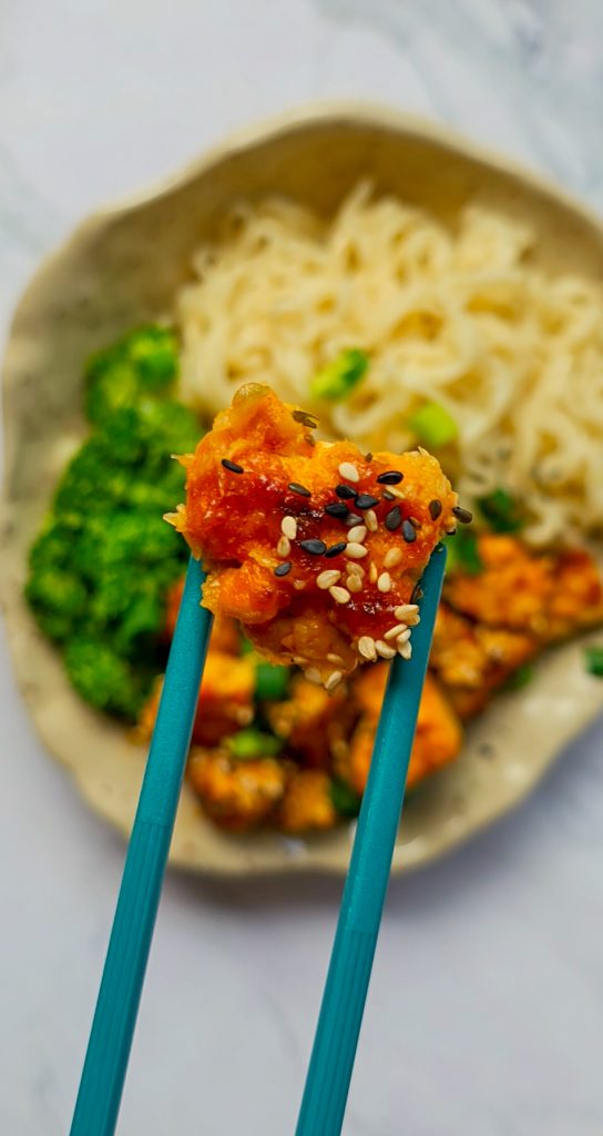 Mit Panade und Sesam ummantelter knuspriger Tofu zwischen zwei Ess-Stäbchen über einem Teller mit Tofu, Mie-Nudeln und Brokkoli.