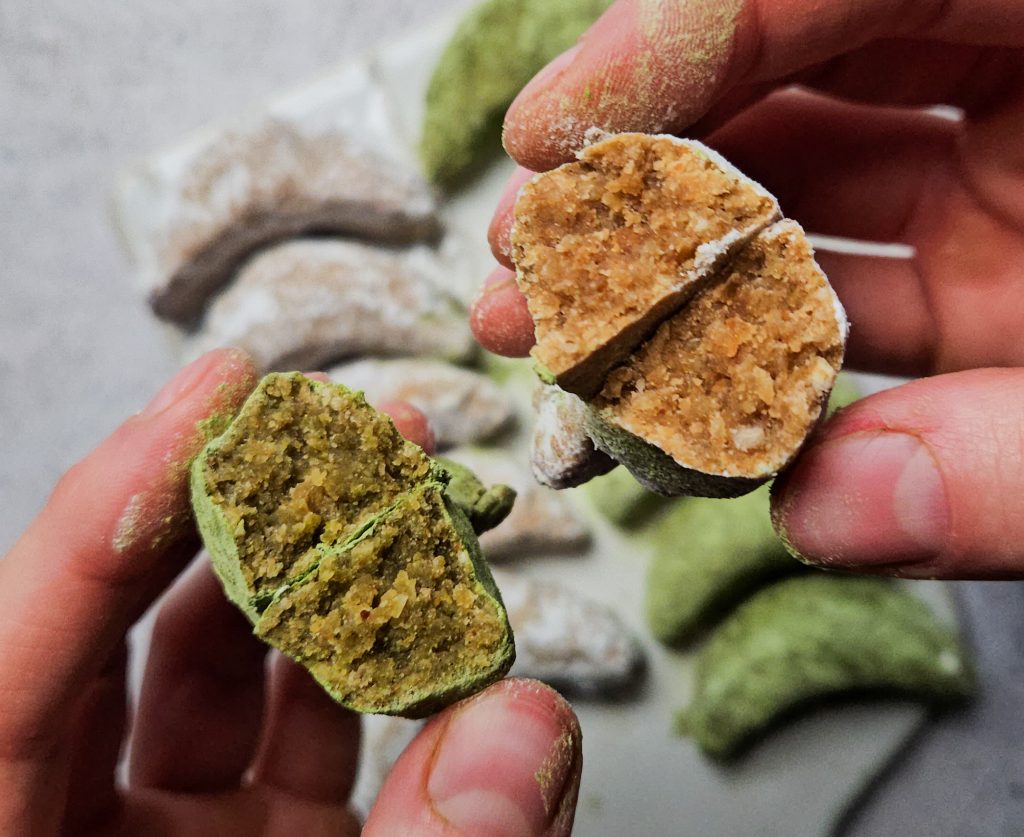 Zwei Hände halten aufgebrochene Vanillekipferl mit grüner Matcha- und heller Vanillefüllung, im Hintergrund liegen weitere Kipferl auf einem Tablett.