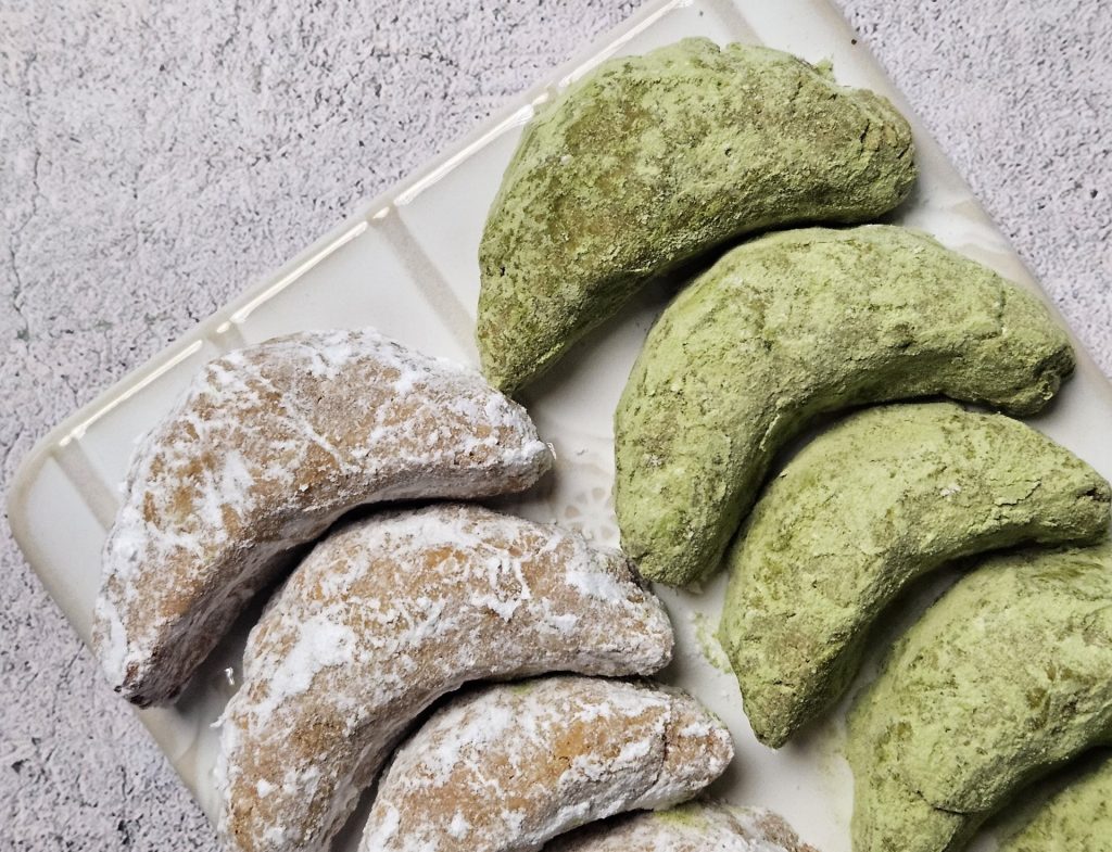 Eine weiße Platte mit veganen Vanillekipferln, zur Hälfte mit Puderzucker bestäubt und zur Hälfte mit Matcha-Pulver umhüllt.
