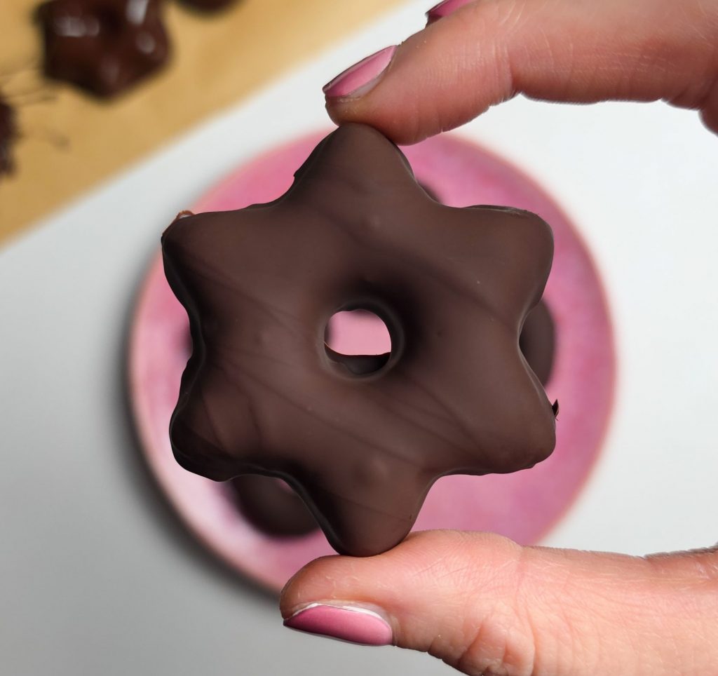 Hand hält einen sternförmigen, mit dunkler Schokolade überzogenen veganen Lebkuchen vor einem rosa Teller.