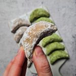 Eine Hand hält ein einzelnes veganes Vanillekipferl mit Puderzucker, im Hintergrund sind weitere Kipferl auf einer Platte zu sehen.