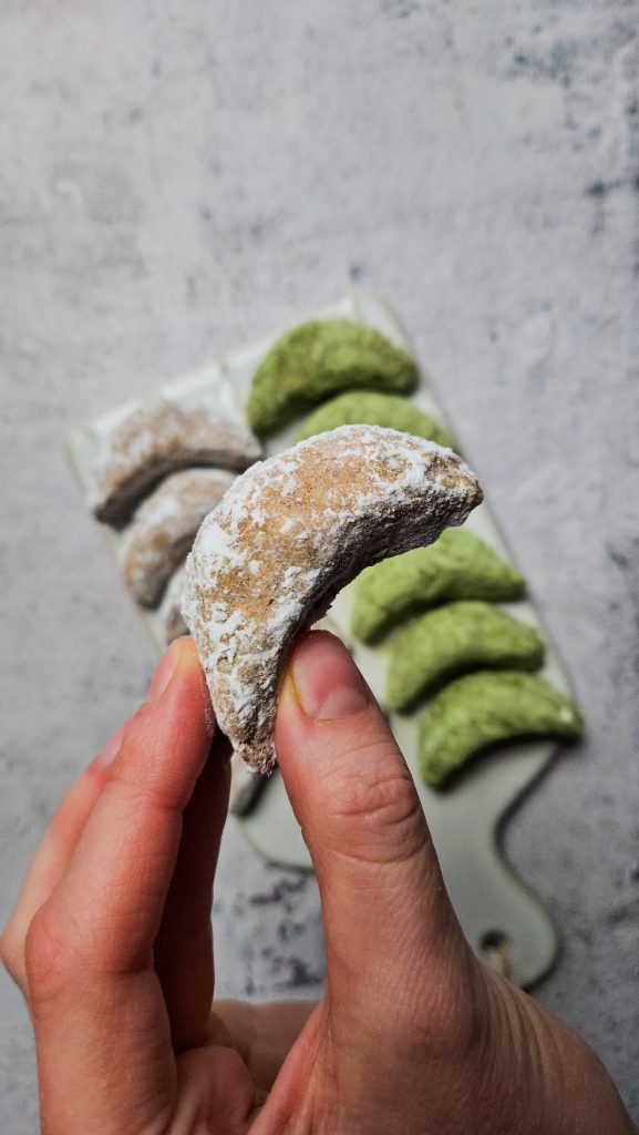 Eine Hand hält ein einzelnes veganes Vanillekipferl mit Puderzucker, im Hintergrund sind weitere Kipferl auf einer Platte zu sehen.