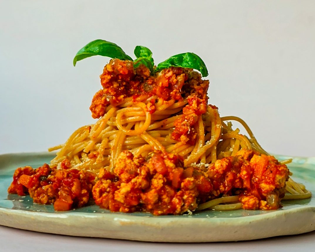 Vegane Bolognese mit Basilikum angerichtet auf einem blau-grünen Teller vor weißem Hintergrund.