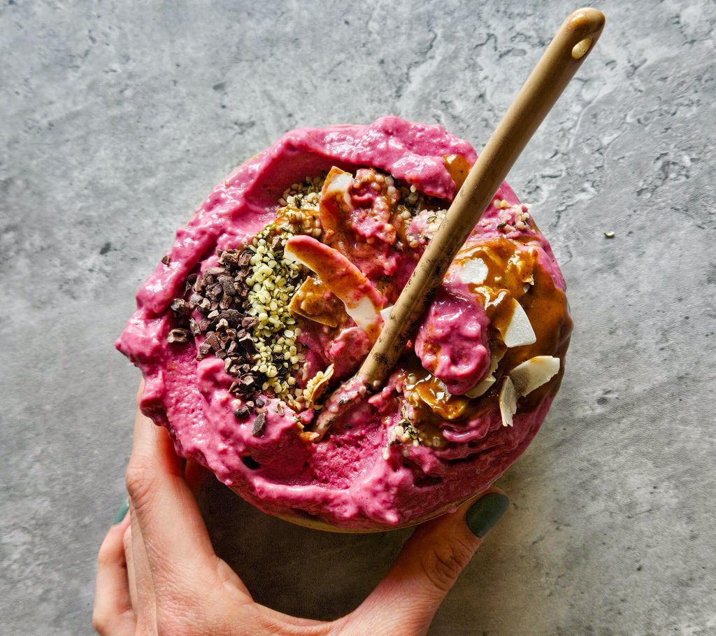 Eine Hand hält eine pinke gefrorene Smoothiebowl in einer Schale, die mit Kakaonibs, Hanfsamen, Kokoschips und Mandelmus getoppt ist. In der Schale befindet sich ein Löffel