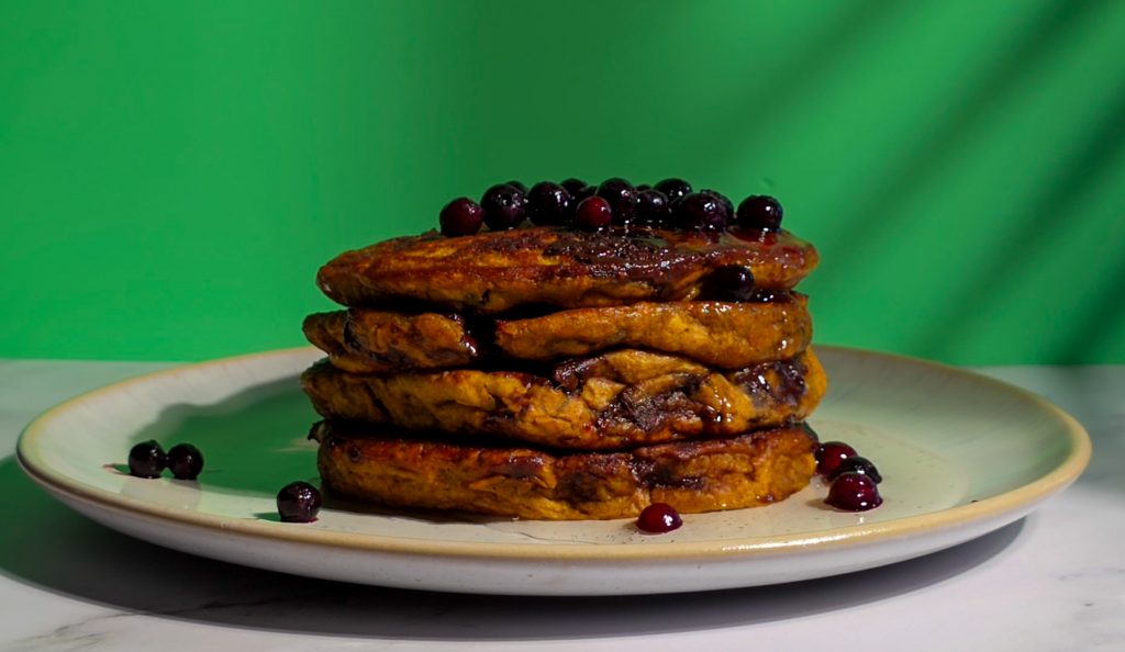 Saftige vegane Kürbis-Pancakes mit Schokostückchen, gestapelt auf einem Teller, getoppt mit Blaubeeren und Ahornsirup vor grünem Hintergrund.