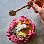 Pinke gefrorene Smoothiebowl in einer Schale, getoppt mit Kakaonibs, Hanfsamen, Kokoschips und Mandelmus, daneben eine Hand mit Löffel