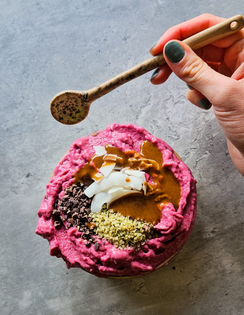 Pinke gefrorene Smoothiebowl in einer Schale, getoppt mit Kakaonibs, Hanfsamen, Kokoschips und Mandelmus, daneben eine Hand mit Löffel