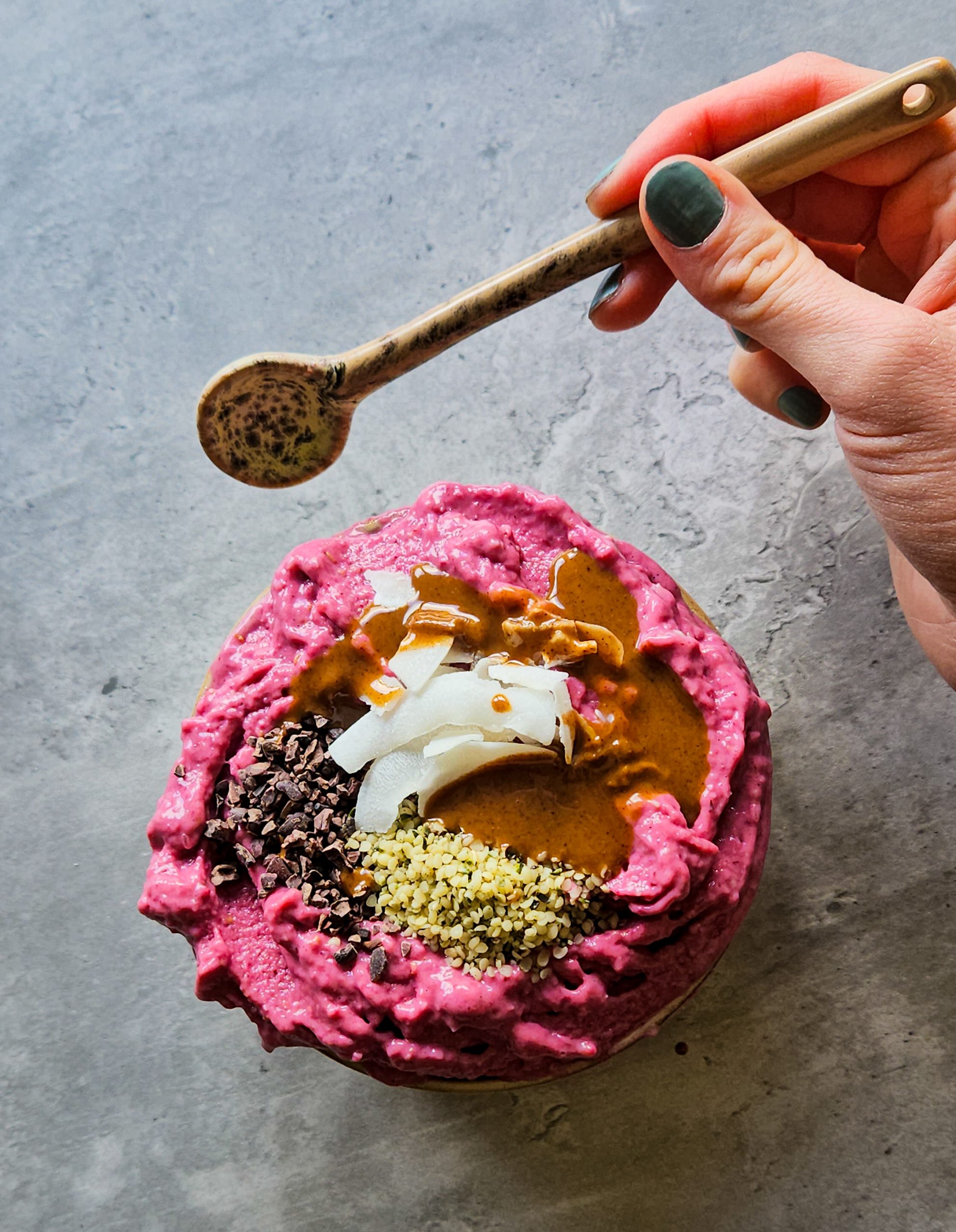 Pinke gefrorene Smoothiebowl in einer Schale, getoppt mit Kakaonibs, Hanfsamen, Kokoschips und Mandelmus, daneben eine Hand mit Löffel