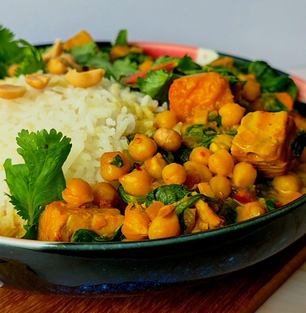 Veganes Kürbis Curry mit Kichererbsen, Spinat, Paprika und Tempeh, serviert mit Reis und garniert mit frischem Koriander und Erdnüssen in einer schwarzen Schale.