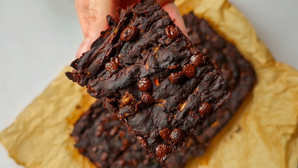 Hand hält ein Stück saftigen veganen Zucchini-Brownie mit geschmolzenen Schokostückchen.