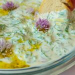 Eine Scheibe Baguette, die in einen Kräuterquark verziert mit Olivenöl und Schnittlauchblüten gedippt wird.