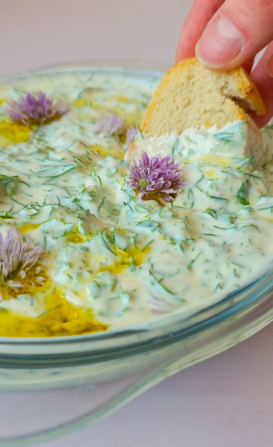 Eine Scheibe Baguette, die in einen Kräuterquark verziert mit Olivenöl und Schnittlauchblüten gedippt wird.