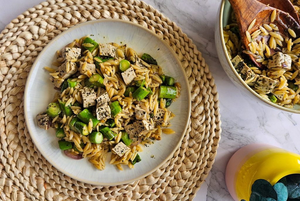 Salat mit Orzo, Spargel, Walnüssen, veganem Feta und Dressing auf einem beigen Teller neben der gefüllten Salatschüssel