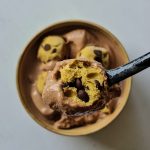 Auf einem schwarzen Löffel befindet sich Schokoladeneis und ein halbes Cookie Dough Bällchen, dahinter sieht man eine ganze Schale mit Schokoladeneis und Cookie Dough.