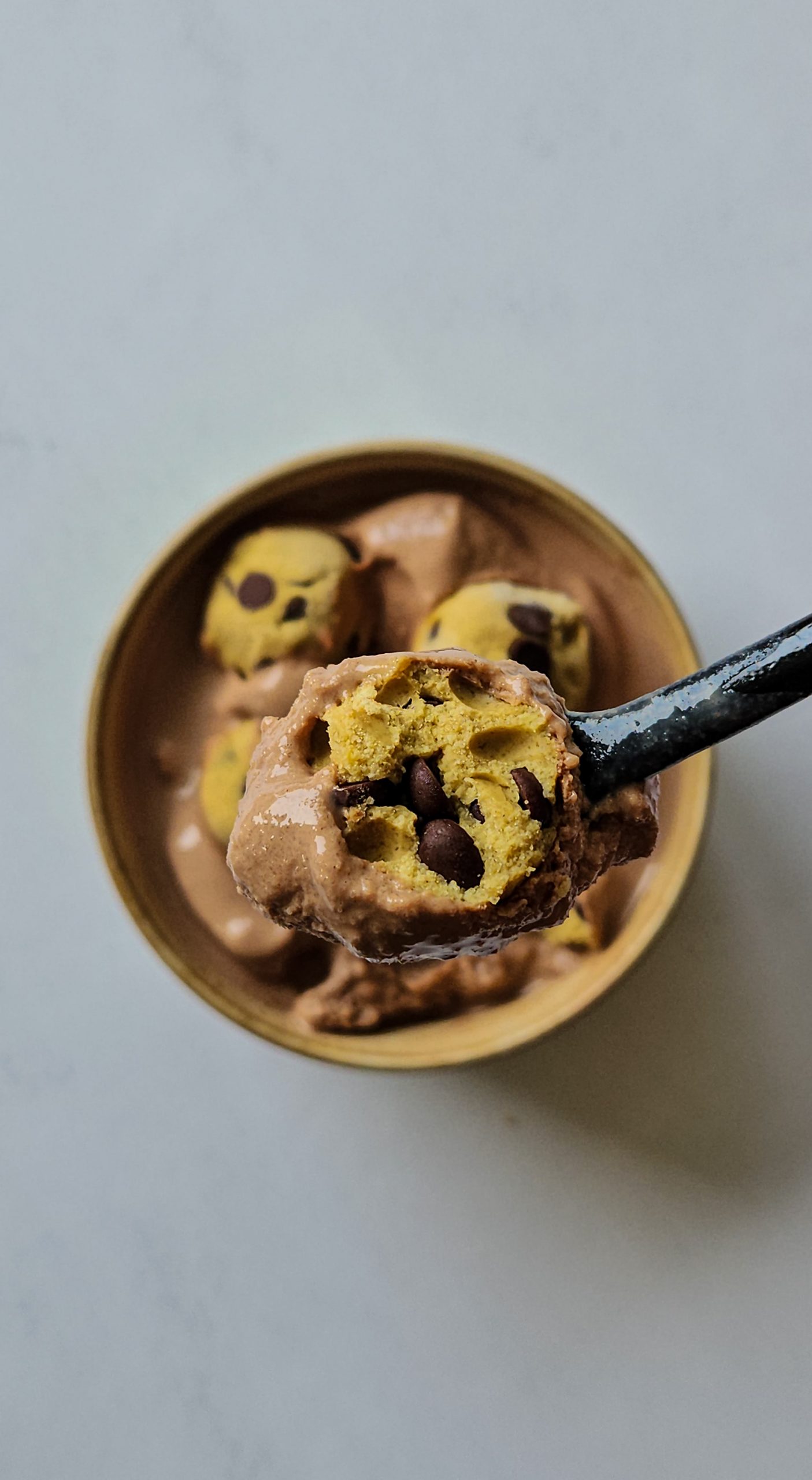 Auf einem schwarzen Löffel befindet sich Schokoladeneis und ein halbes Cookie Dough Bällchen, dahinter sieht man eine ganze Schale mit Schokoladeneis und Cookie Dough.